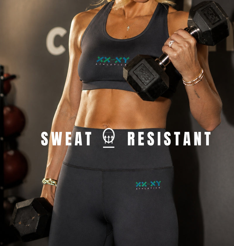 A woman in a gym wearing a dark gray sports bra and matching high-waisted leggings with the XX-XY Athletic logo, holding dumbbells. Large white text overlays the center reading 'SWEAT RESISTANT'.