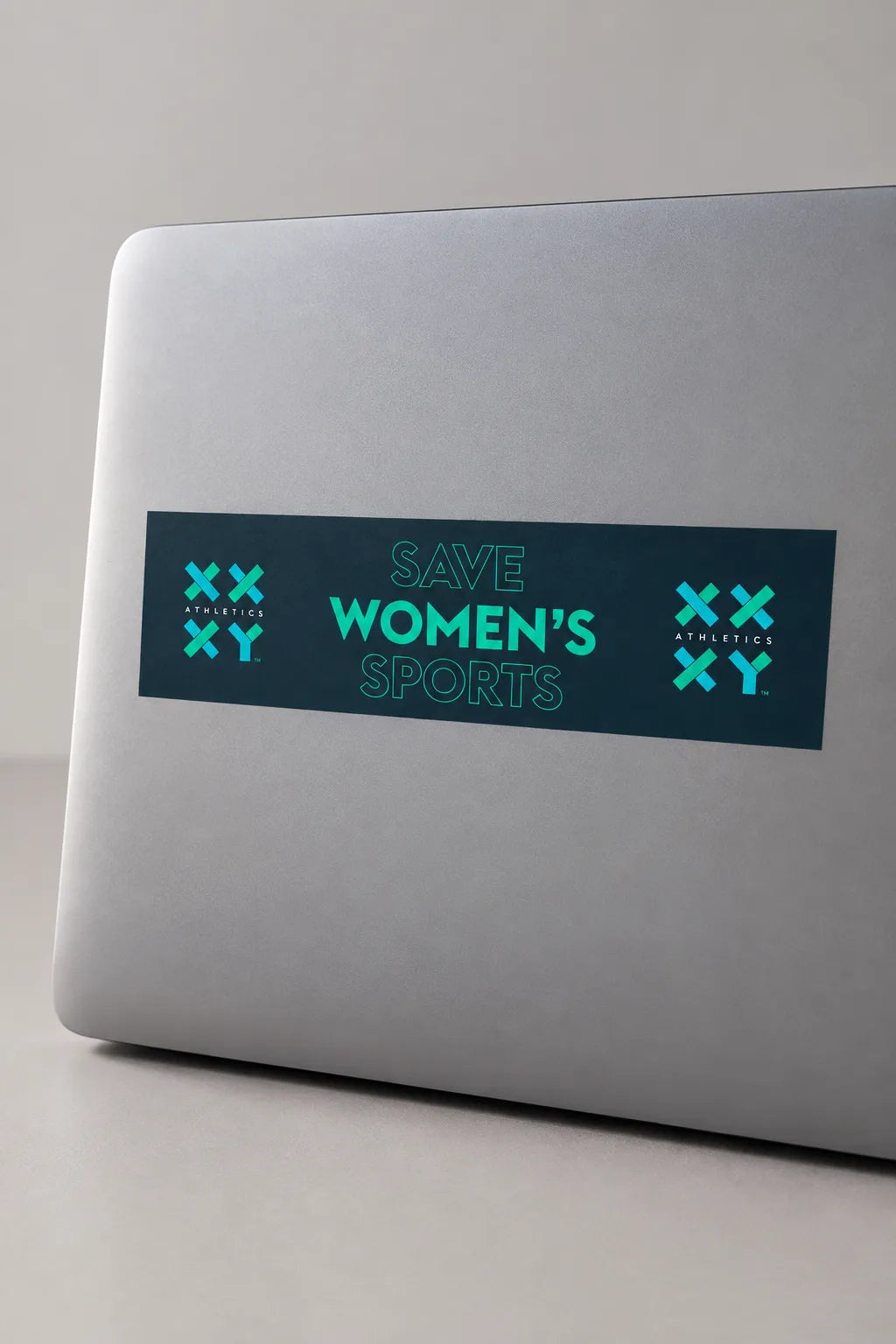 A rectangular bumper sticker with the text 'SAVE WOMEN'S SPORTS' in teal on a dark teal background, displayed on a gray laptop.