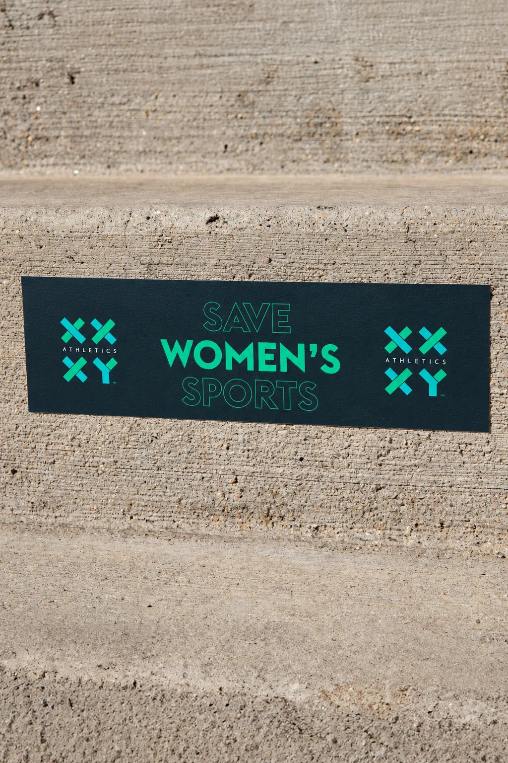 A rectangular black bumper sticker with the text 'SAVE WOMEN'S SPORTS' in bright green, displayed on concrete steps. The sticker features the 'XXY Athletics' logo on both sides.