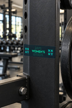 A rectangular bumper sticker with the text 'SAVE WOMEN'S SPORTS' in teal on a dark teal background, displayed on a piece of gym equiptment.