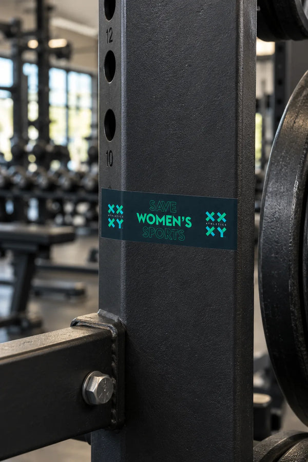 A rectangular bumper sticker with the text 'SAVE WOMEN'S SPORTS' in teal on a dark teal background, displayed on a piece of gym equiptment.