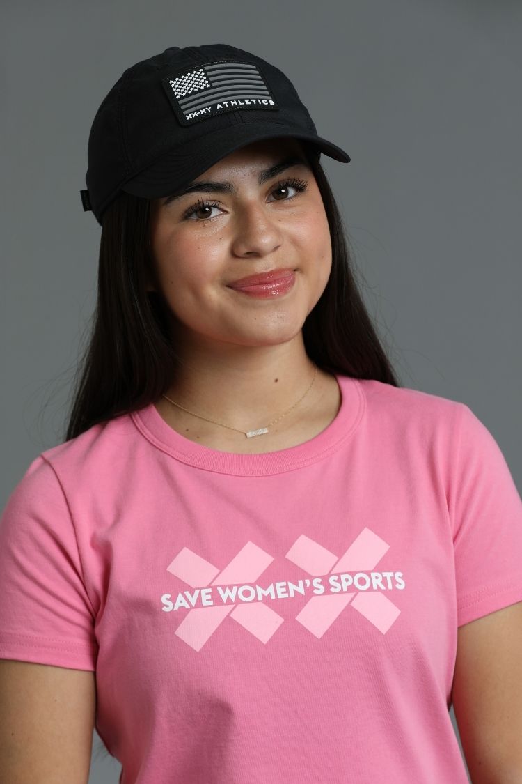 Person wearing a pink 'Save Women's Sports' t-shirt and black cap with an American flag design. || Pink