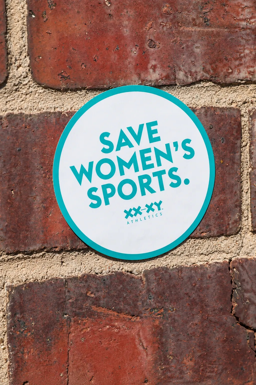A round white sticker with a teal border and the text 'SAVE WOMEN'S SPORTS.' and 'XXY ATHLETICS' in teal, shown against a red-brown brick wall.