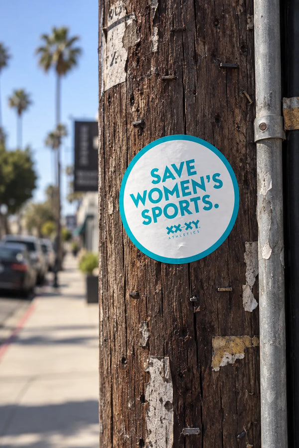 Round Save Women’s Sports XX-XY Athletics sticker displayed on a wooden telephone pole.