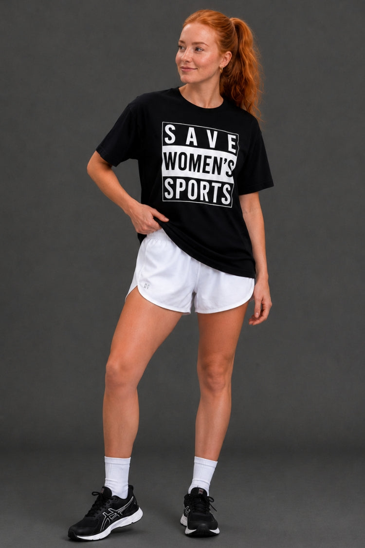 A woman modeling a black graphic t-shirt with white text that reads 'SAVE WOMEN'S SPORTS', paired with white athletic shorts, white socks, and black sneakers, against a dark gray studio background. || Black