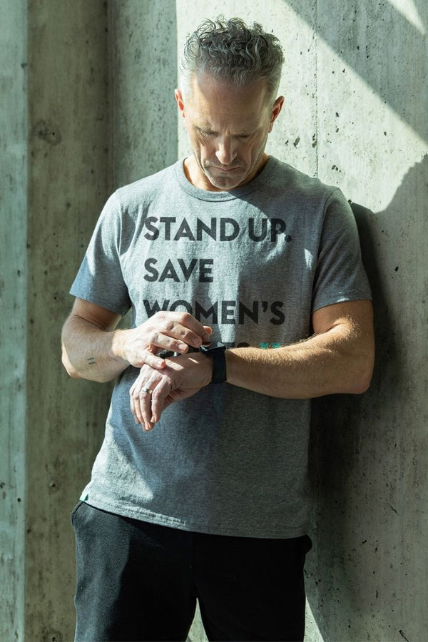 Man wearing a t-shirt with 'STAND UP. SAVE WOMEN'S' text against a concrete wall. || heather gray