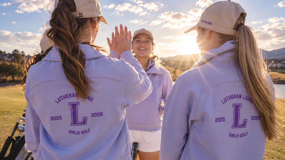 Three young women on a golf course at sunset wearing matching light lavender-purple hooded jackets with purple 'Lutheran Lions 2025 Girls Golf' back graphics and beige baseball caps, high-fiving in the foreground.