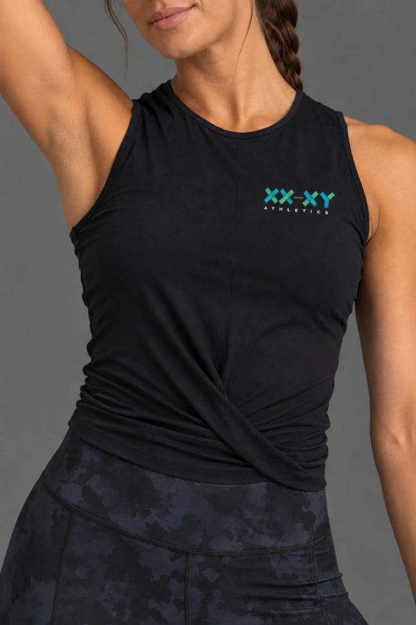 Close-up shot of a woman modeling a black sleeveless athletic crop top with a twisted front detail and a small XX-XY Athletics logo on the chest, paired with camo leggings against a solid gray studio background. || Black