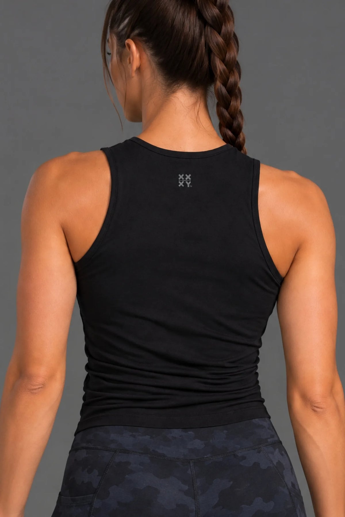 Back shot of a woman modeling a black sleeveless athletic crop top with a small XX-XY Athletics logo on upper back, paired with camo leggings against a solid gray studio background. || Black