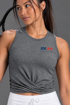 A woman modeling a heather gray sleeveless athletic crop top with a twisted front detail and a small XX-XY Athletics logo on the chest, paired with a white tennis skirt against a solid gray studio background. || Dark Heather Gray