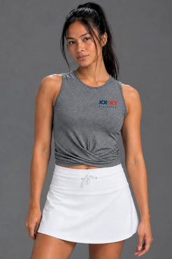A woman modeling a heather gray sleeveless athletic crop top with a twisted front detail and a small XX-XY Athletics logo on the chest, paired with a white tennis skirt against a solid gray studio background. || Dark Heather Gray