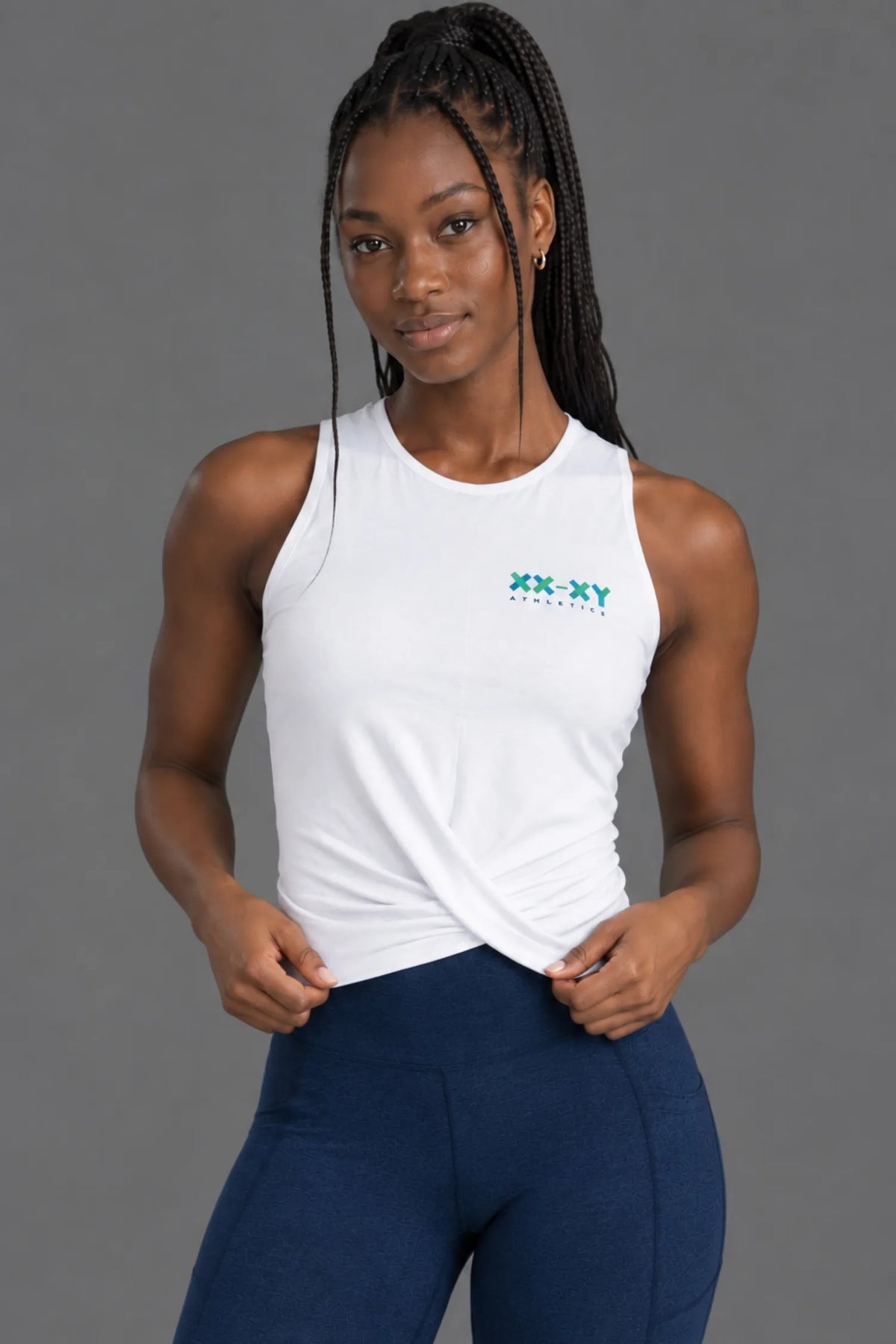 Female model wearing a white XX-XY Athletics twist-front tank top with navy leggings. || White