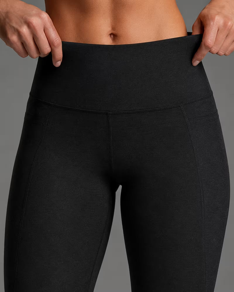 Close-up of a model wearing black high-waisted leggings, pulling the waistband outward with both hands to demonstrate the stretch and fit. The background is a solid neutral gray.