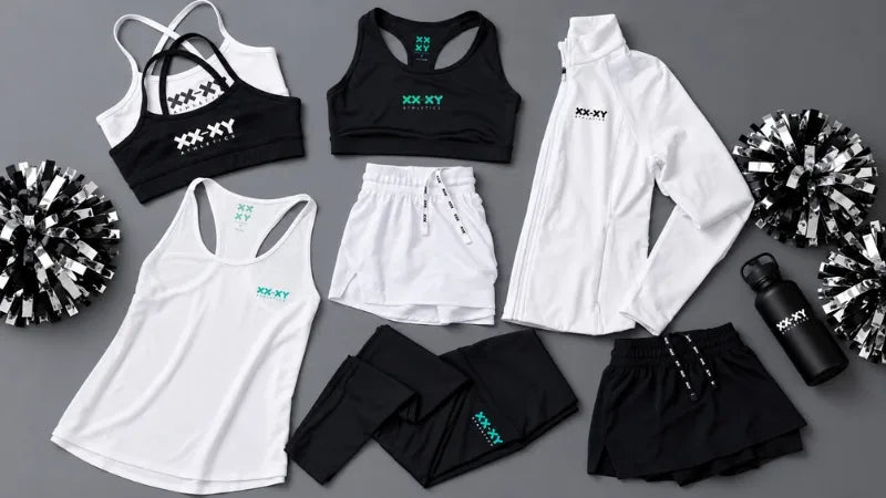 A coordinated activewear set laid out on a gray background, including a white racerback tank, black sports bra, white shorts, black leggings, a white zip jacket, black shorts, a black water bottle, and black-and-white pom-poms. The apparel features the XXX-XY brand name in white and teal.