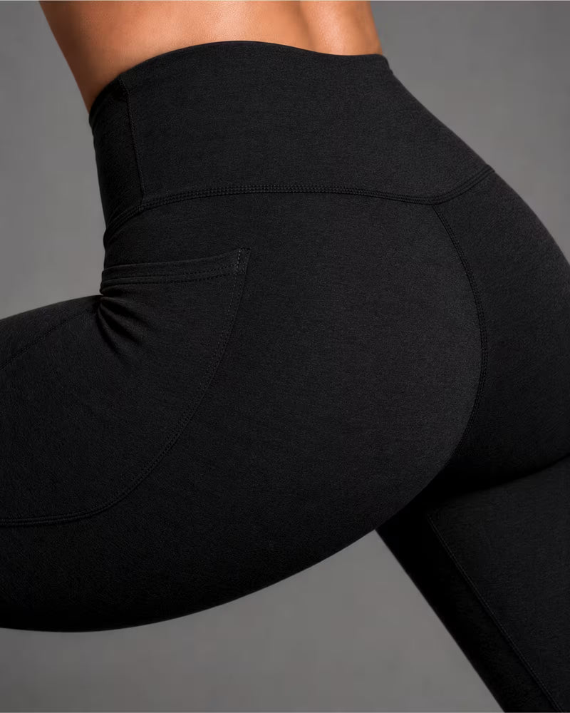 Close-up of a model wearing black high-waisted leggings, showing the side pocket and waistband against a neutral gray background.