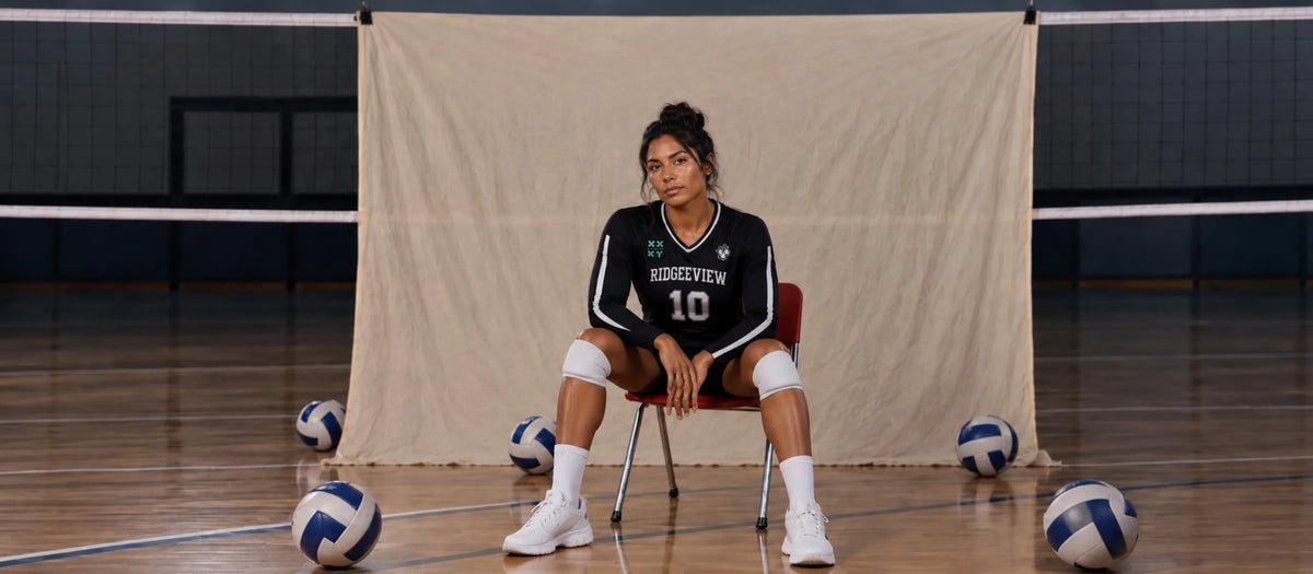 A woman sits on a red chair in an indoor volleyball court, wearing a black and white FDU Eagles volleyball jersey with the number 10. Volleyballs are scattered on the wooden floor around her, with a beige fabric backdrop behind her.