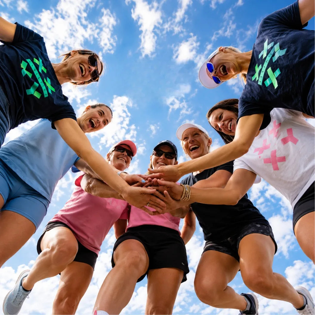 Six women in athletic wear jump together in a circle, stacking their hands in the center, against a bright blue sky with white clouds. They are wearing matching dark athletic shirts with colorful geometric logos (green, pink, and teal) and shorts in various colors.