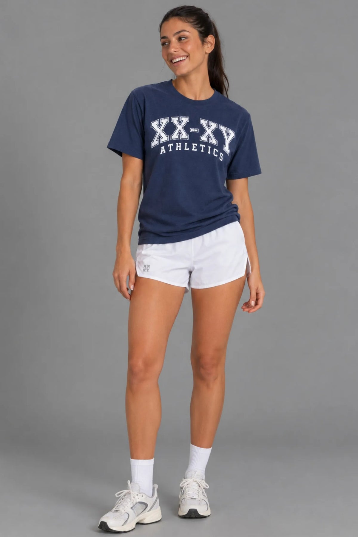 Female model laughing while wearing the XX-XY Athletics Varsity Logo Tee, white running shorts, crew socks, and sneakers