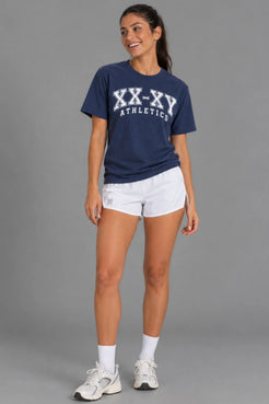 Female model laughing while wearing the XX-XY Athletics Varsity Logo Tee, white running shorts, crew socks, and sneakers || Navy