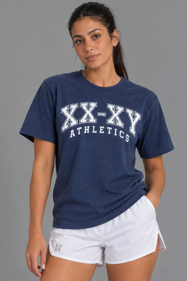 Female model wearing the XX-XY Athletics Varsity Logo Tee with distressed white collegiate lettering, paired with white athletic shorts against a gray background || Navy