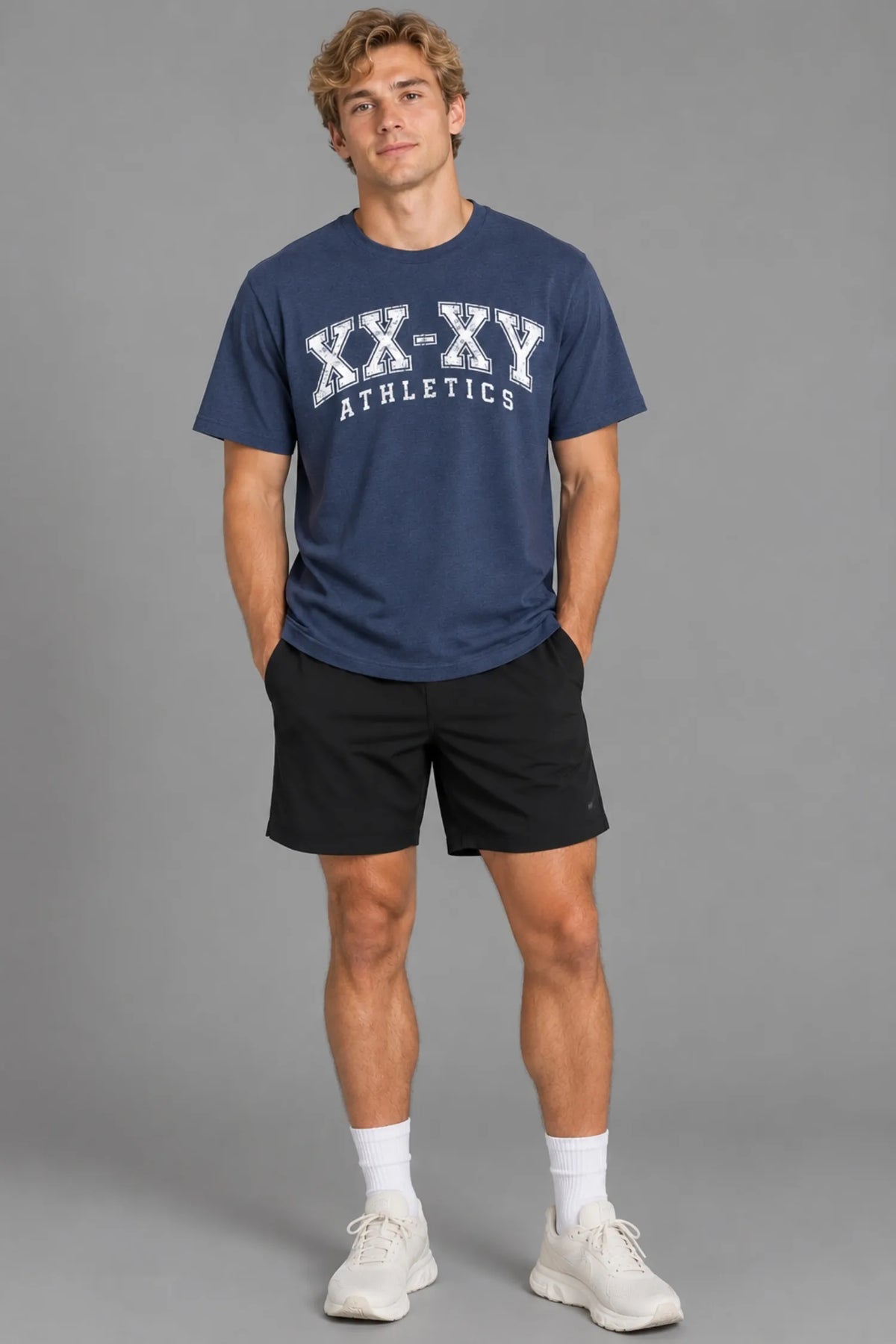 Male model wearing the XX-XY Athletics Varsity Logo Tee with black shorts, white socks, and sneakers, standing against a gray background