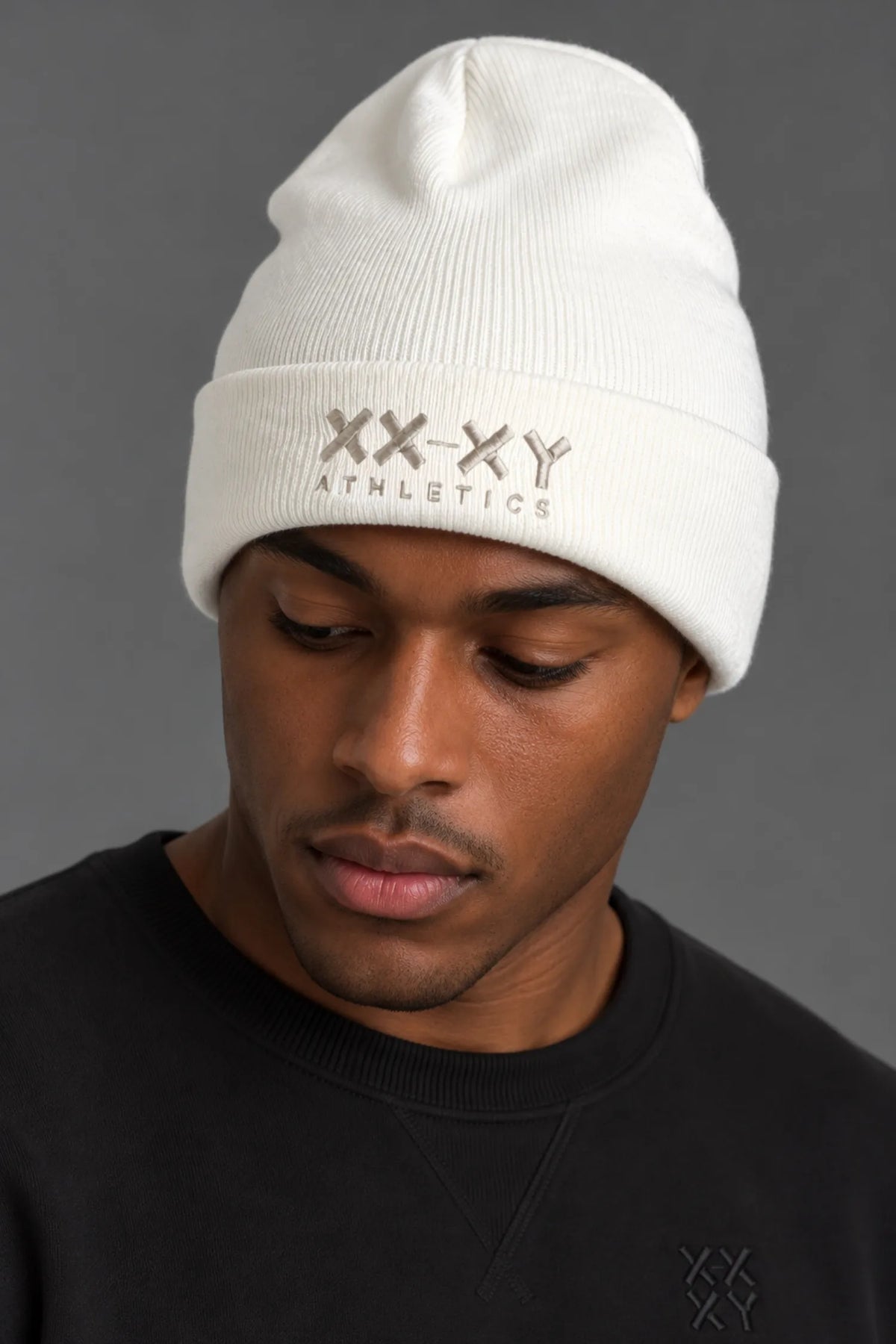 Black male model wearing white XX-XY Athletics embroidered beanie and black crewneck sweatshirt against a gray background