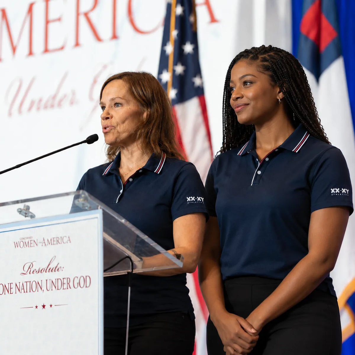 Two women wearing navy XX-XY Athletics polos stand side by side at a podium, with one speaking into a microphone and the other smiling softly against a patriotic backdrop with American flags.