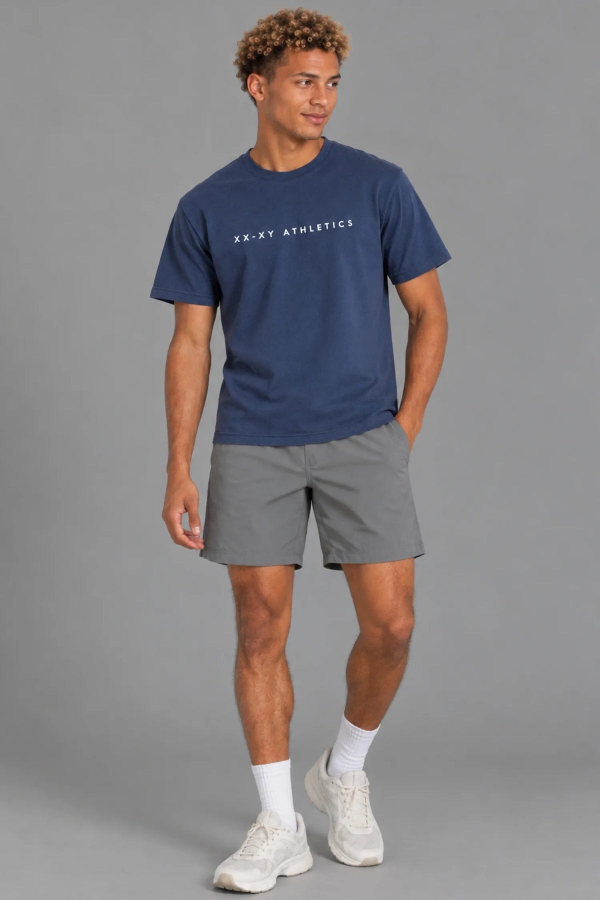 Male model wearing the XX-XY Athletics Wordmark Logo Tee with gray shorts, white socks, and sneakers, standing against a gray background || Navy