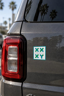 A square sticker with a green and white XXYY Athletics logo applied to the rear quarter panel of a dark gray vehicle, with palm trees and a sunny outdoor background reflected on the car. || White