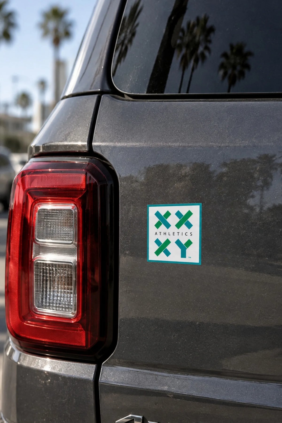 A square sticker with a green and white XXYY Athletics logo applied to the rear quarter panel of a dark gray vehicle, with palm trees and a sunny outdoor background reflected on the car. || White
