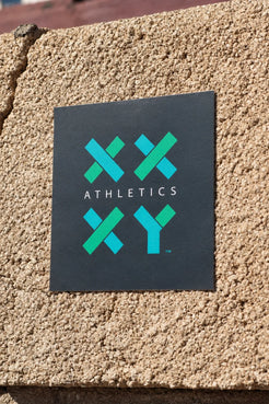 XX-XY Athletics logo sticker displayed on a textured beige stucco wall. || Dark