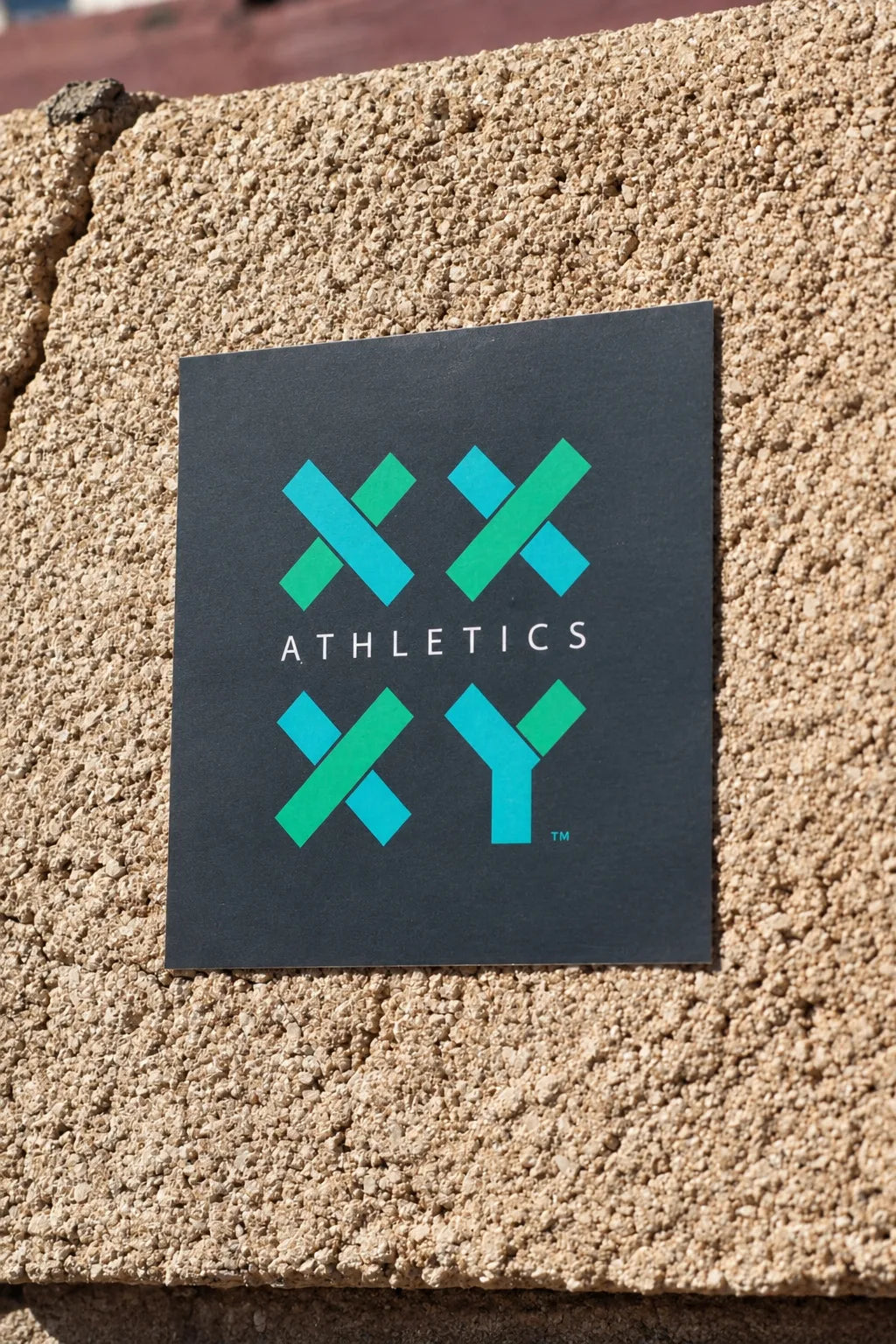 XX-XY Athletics logo sticker displayed on a textured beige stucco wall. || Dark
