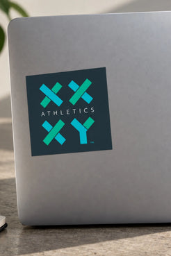 A square sticker with the text 'ATHLETICS' and a teal and blue geometric logo is applied to the back of a silver laptop. The laptop is resting on a stone or concrete surface with a blurred background. || Dark