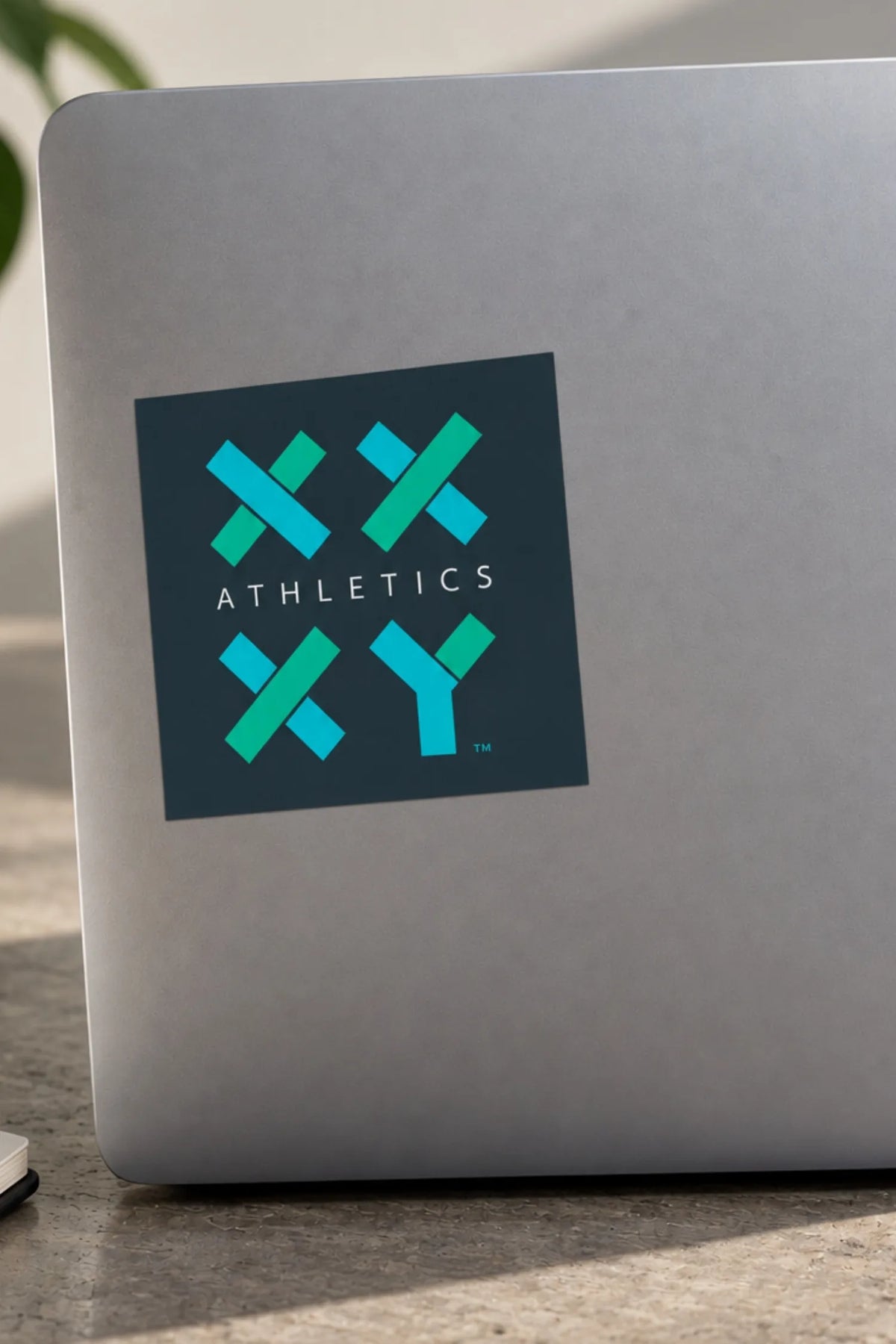 A square sticker with the text 'ATHLETICS' and a teal and blue geometric logo is applied to the back of a silver laptop. The laptop is resting on a stone or concrete surface with a blurred background. || Dark