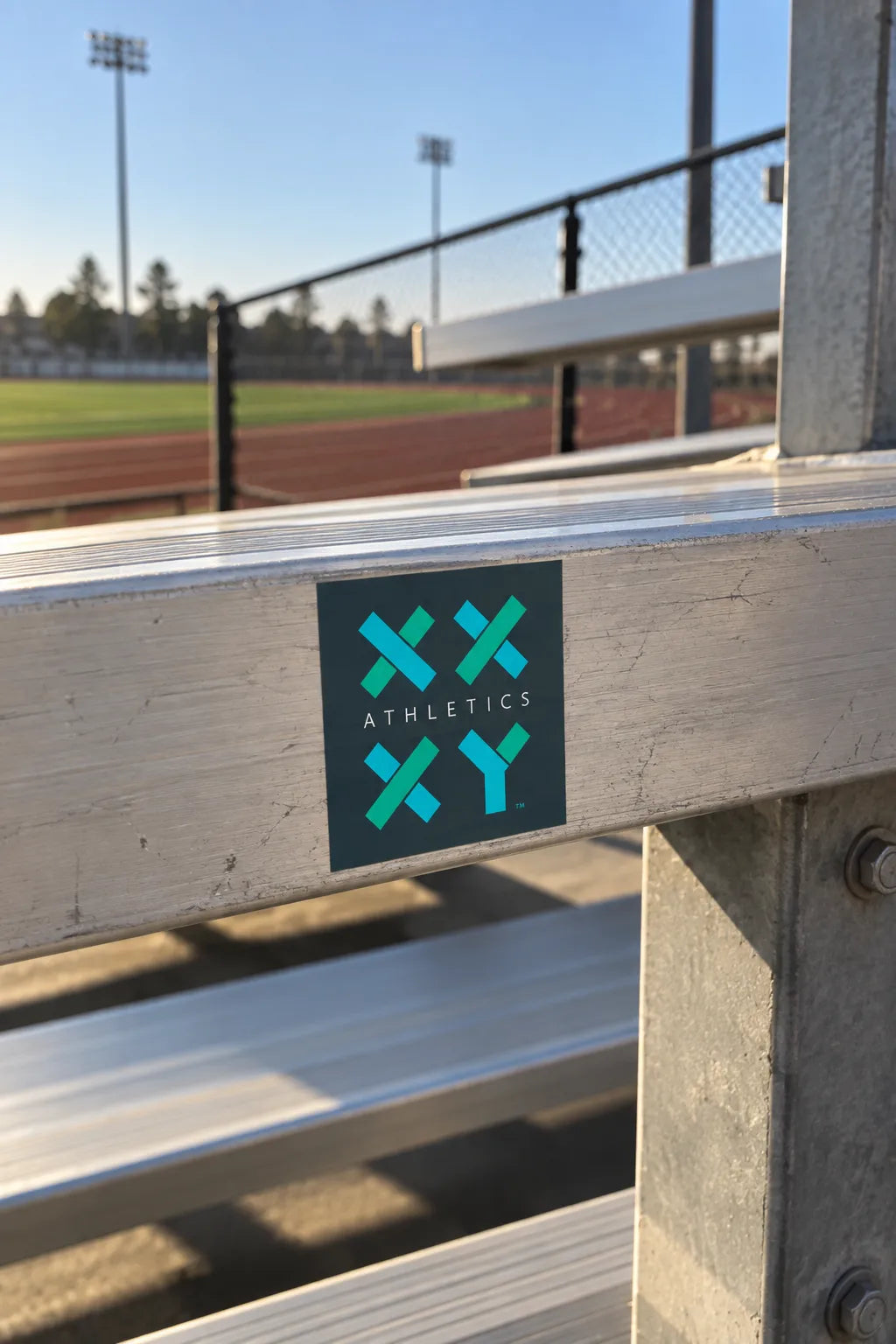 A square black sticker with a teal and green 'XXY Athletics' logo is attached to the metal railing of an outdoor stadium bleacher, with a track and field background visible. || Dark