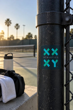 A square sticker with a teal and black grid design and the text 'ATHLETICS XX XY' is shown on a black metal fence post at a tennis court during sunset. A water bottle, towel, and bag are visible in the blurred background. || Dark