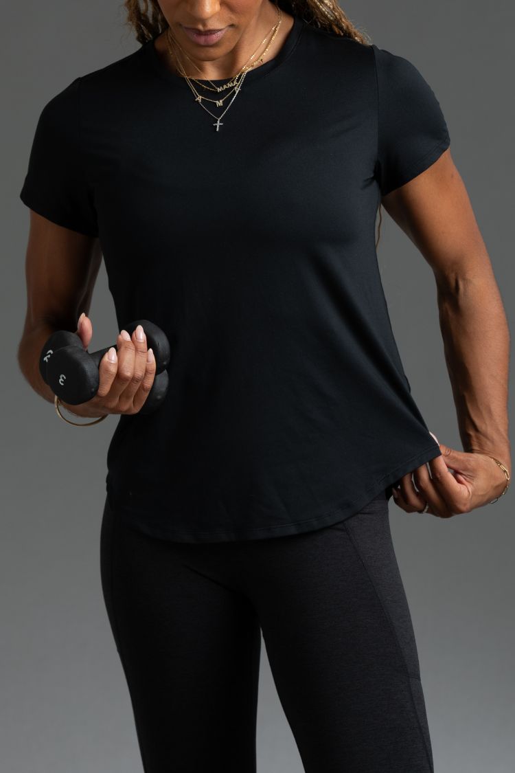 Person wearing a black t-shirt and leggings holding a dumbbell against a gray background || Black