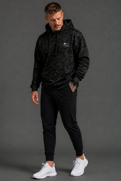 Male model shown from the front wearing a black camo hoodie, black joggers, and white sneakers against a dark gray studio background. || Black Camo