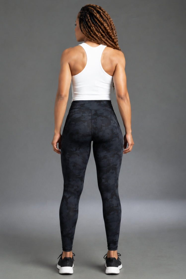 Rear view of a woman wearing high-waisted dark navy and black camouflage leggings with a white tank top, standing against a gray studio background.