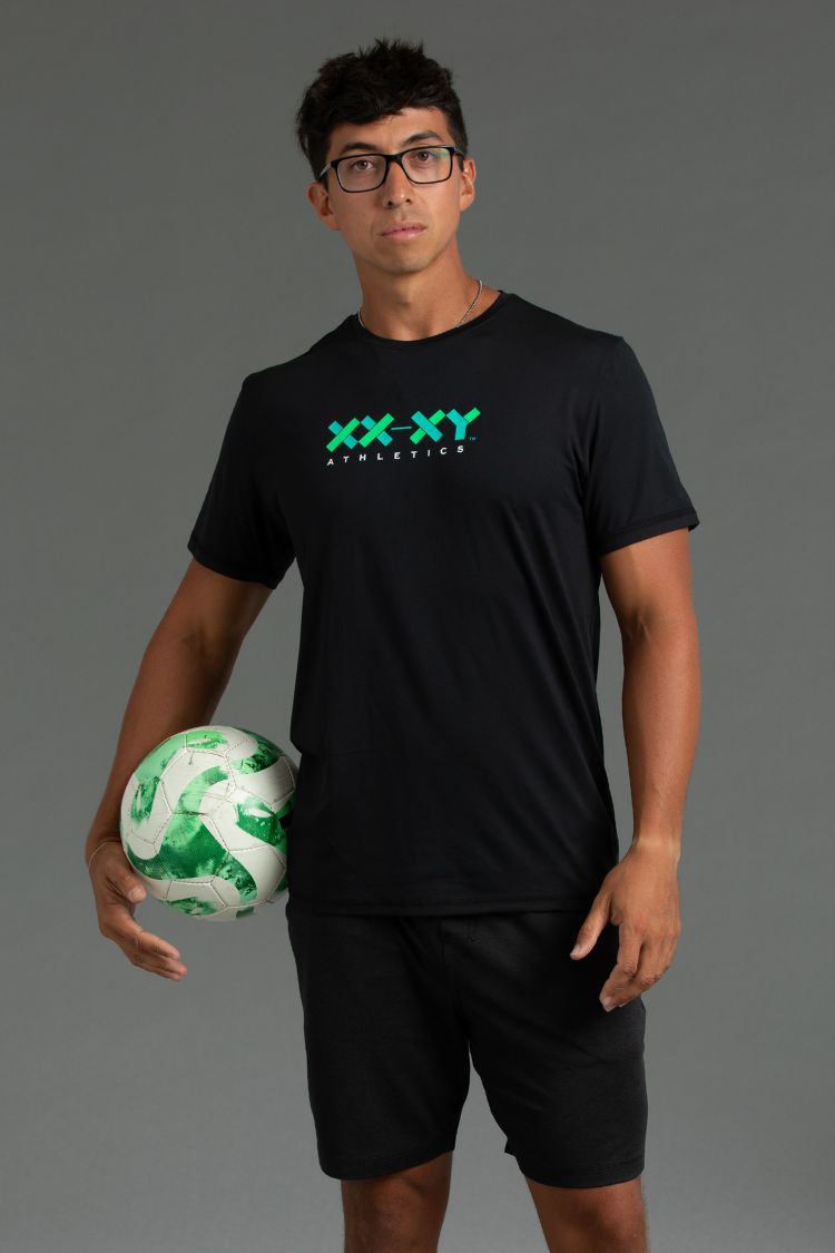 Person wearing a black t-shirt with 'XXXY Athletics' logo, holding a green and white ball against a gray background