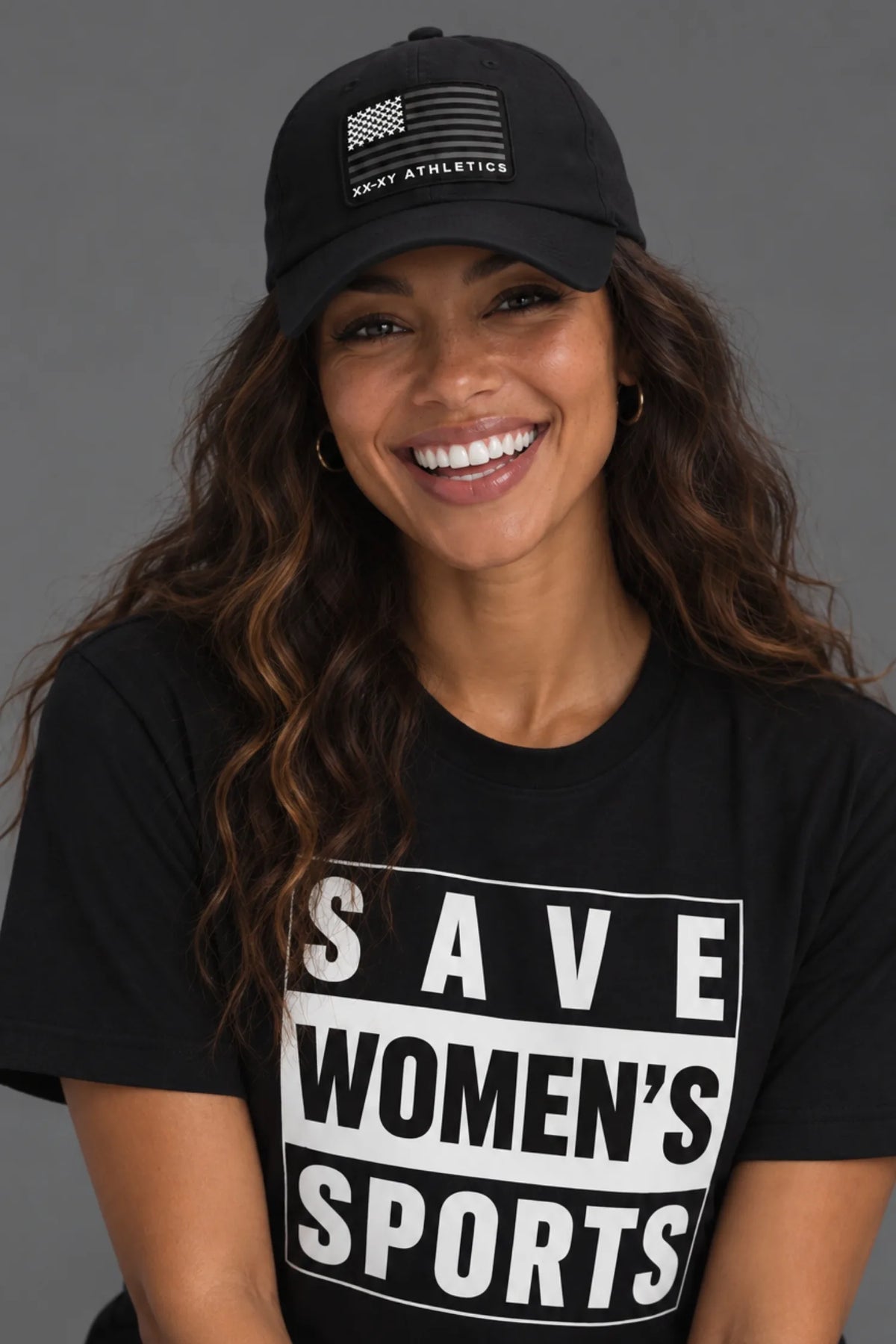 A smiling woman wearing a black baseball cap with a white American flag and 'WOMEN ATHLETICS' text, paired with a black graphic T-shirt that reads 'SAVE WOMEN'S SPORTS' in bold white letters, against a solid gray studio background.