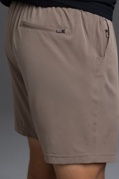 Close-up of a person wearing taupe-brown casual shorts with a zippered pocket and side pockets, paired with a dark navy top against a gray studio background. || Mocha