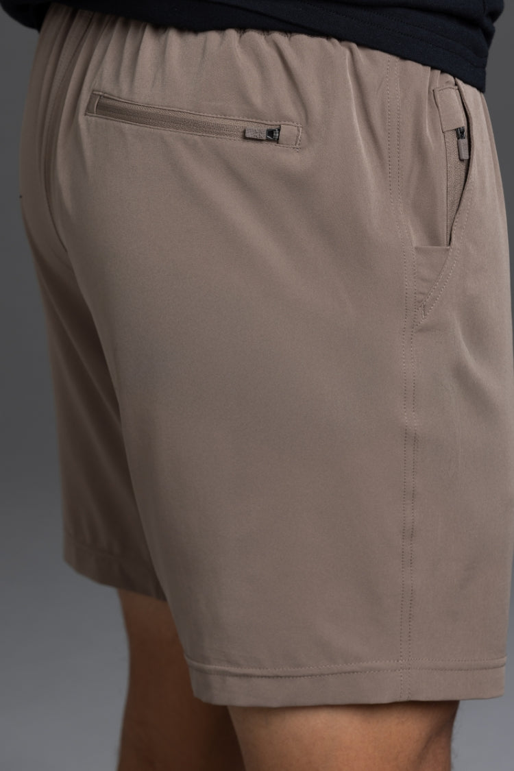 Close-up of a person wearing taupe-brown casual shorts with a zippered pocket and side pockets, paired with a dark navy top against a gray studio background. || Mocha