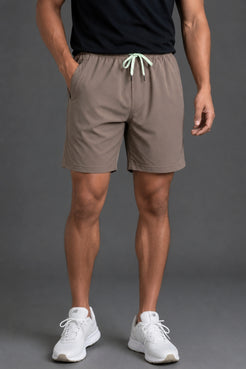 A man wearing taupe-brown athletic shorts with a mint green drawstring, paired with a black t-shirt and white sneakers, shown from the waist down against a gray studio background. || Mocha
