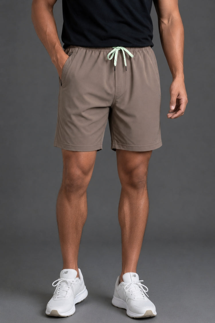 A man wearing taupe-brown athletic shorts with a mint green drawstring, paired with a black t-shirt and white sneakers, shown from the waist down against a gray studio background. || Mocha