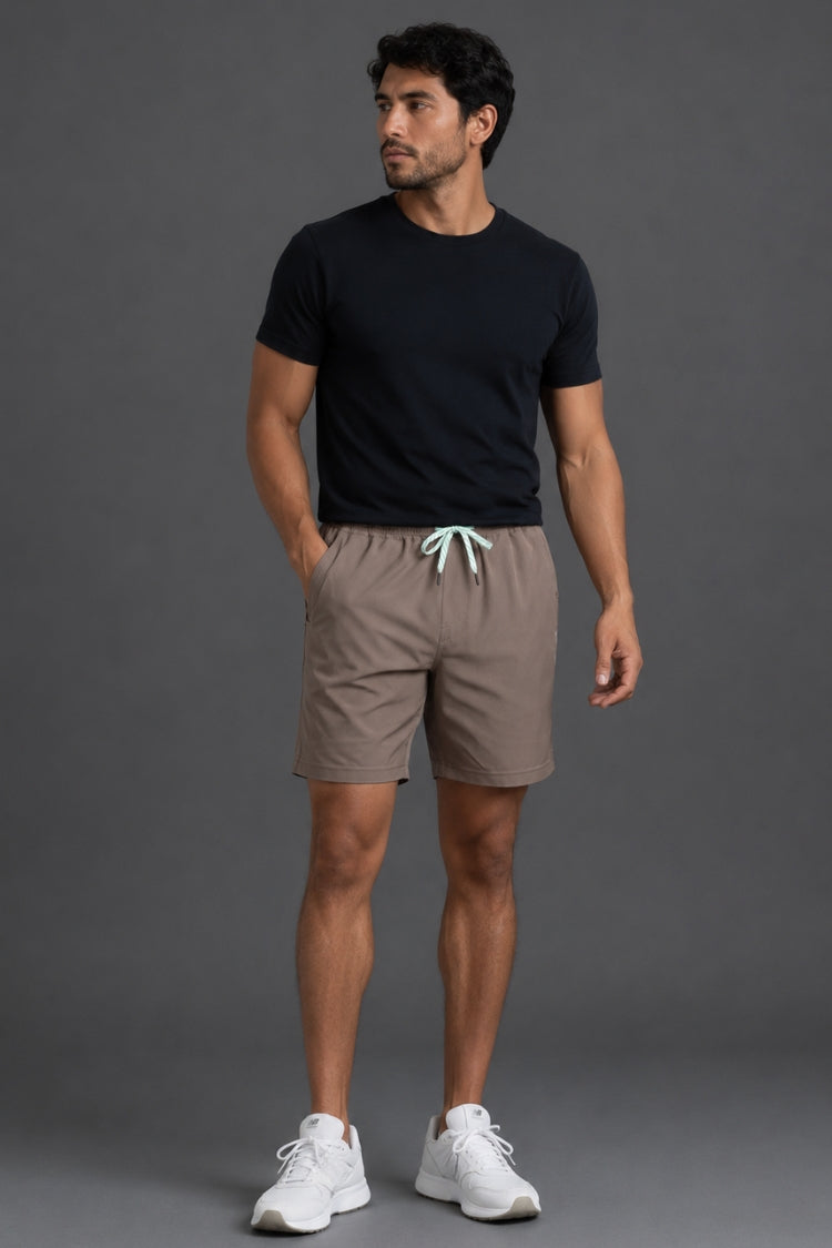 A man wearing taupe-brown athletic shorts with a mint green drawstring, paired with a black t-shirt and white sneakers, shown from the waist down against a gray studio background.
