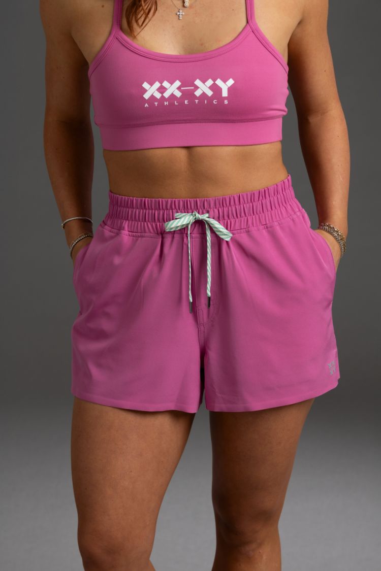 Person wearing pink sports bra and shorts with 'XXXY Athletics' branding on a gray background. || Bright Fuchsia