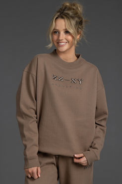 A woman wearing a taupe brown crewneck sweatshirt with a rhinestone-embellished 'XX-XY ATHLETICS' logo on the chest, paired with matching sweatpants, set against a dark gray studio background. || Mocha