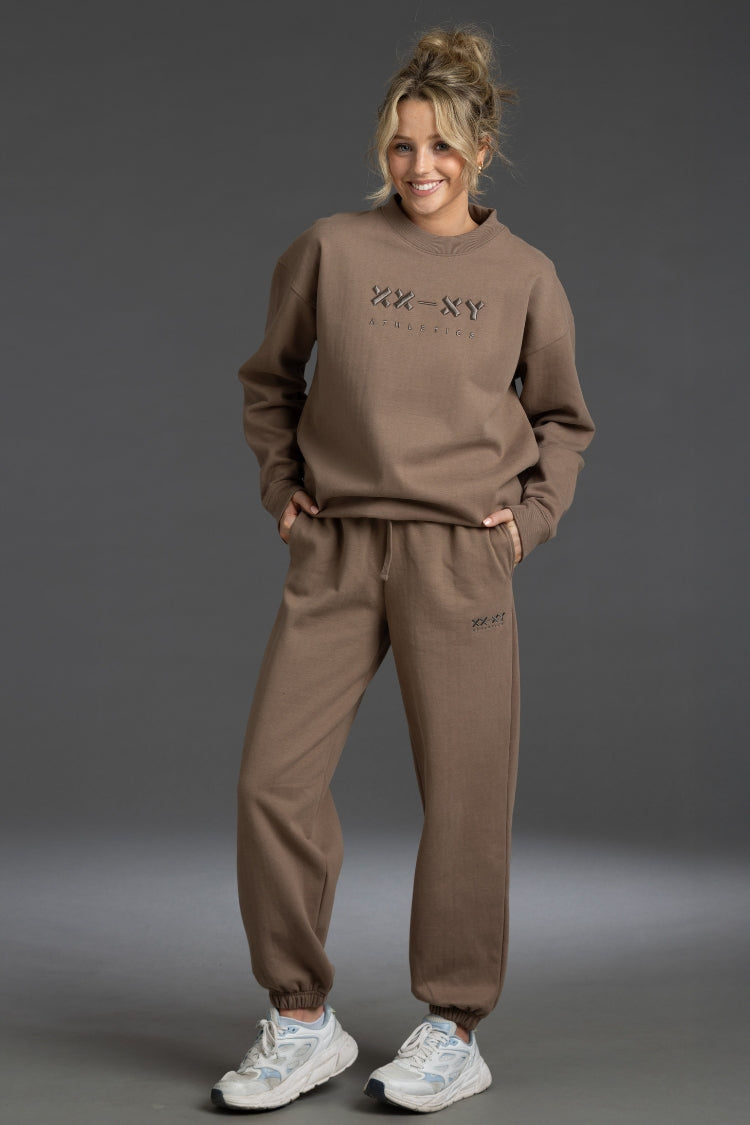 A woman modeling a matching taupe brown sweatshirt and jogger pants set with 'XX-NY' printed on the chest, posed against a dark gray studio background. || Mocha