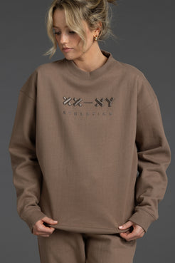 A woman wearing a taupe brown crewneck sweatshirt with a tonal 'XX-XY Aesthetic' logo on the chest, paired with matching sweatpants, set against a dark gray studio background. || Mocha